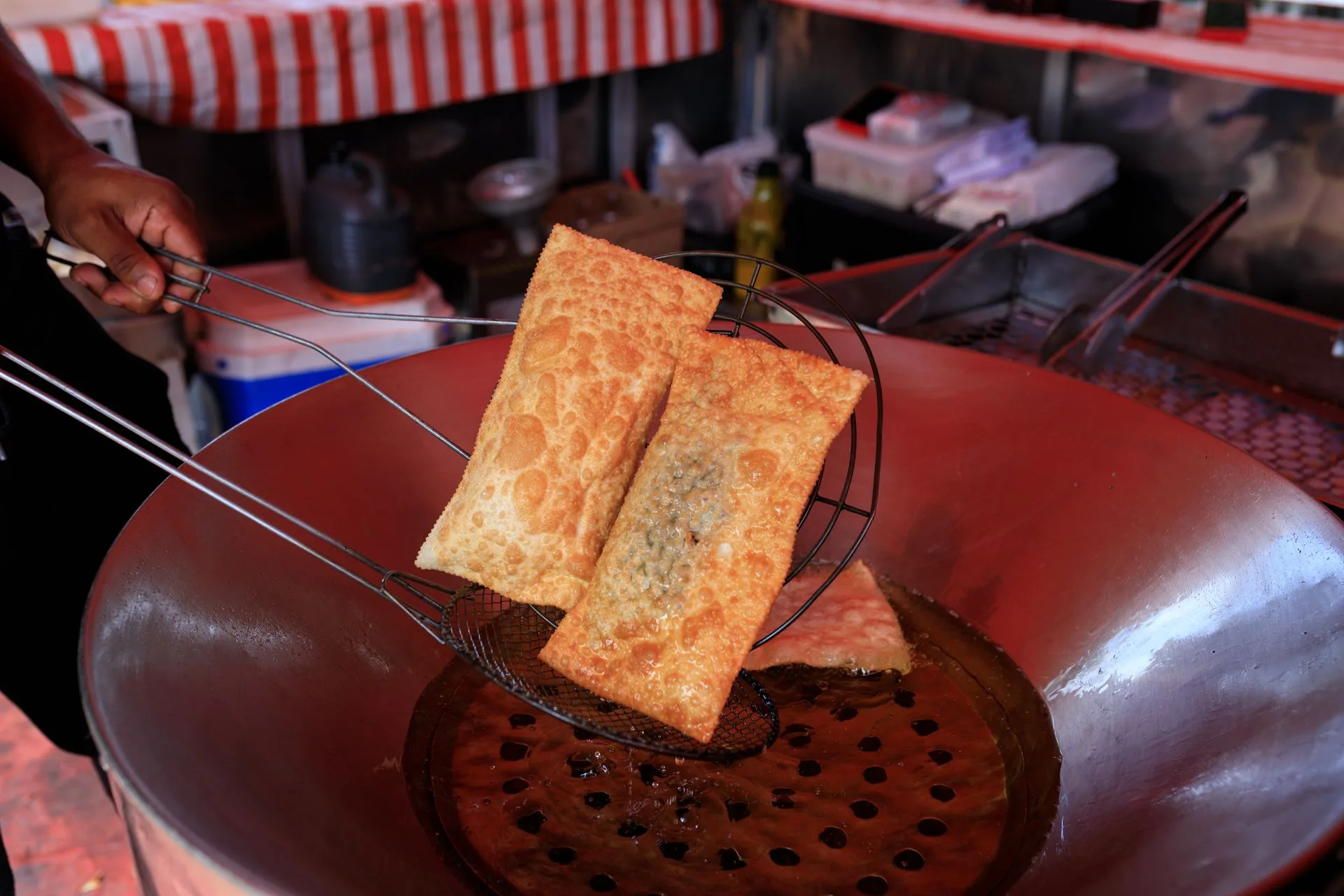 Mão segurando a escumadeira com dois pastéis dourados saindo do tacho de óleo numa barraca de feira