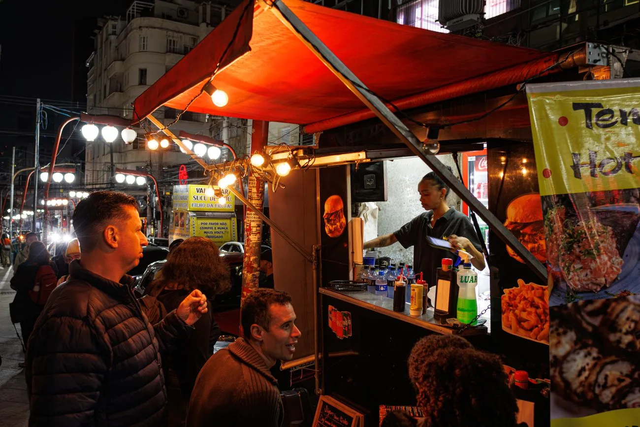 Barraca de lanches numa calçada movimentada de São Paulo à noite, com clientes esperando na frente do balcão iluminado