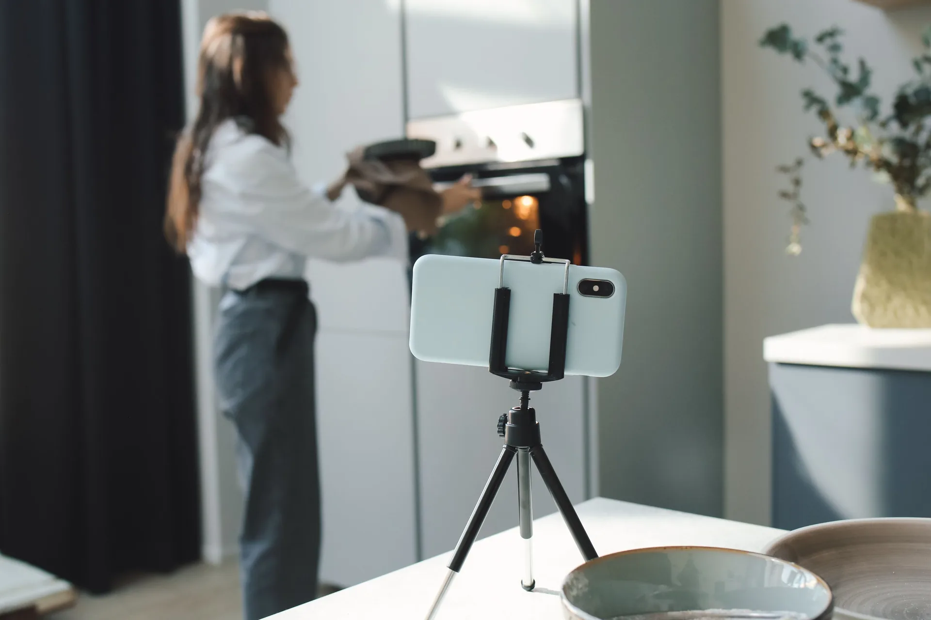 Celular preso num tripé sobre a bancada, gravando uma mulher que tira uma forma do forno ao fundo