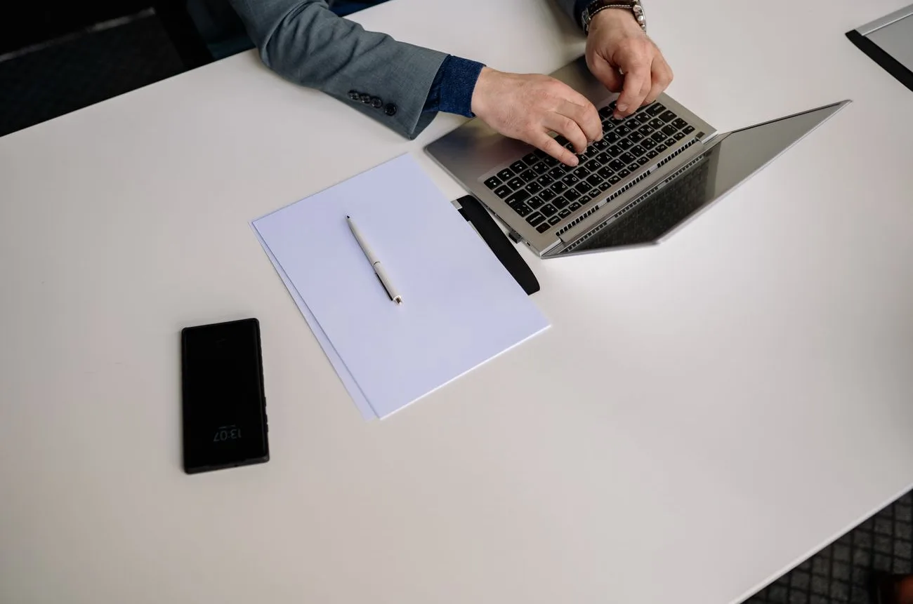 Empresário digitando no notebook com o celular ao lado, em uma mesa de escritório organizada