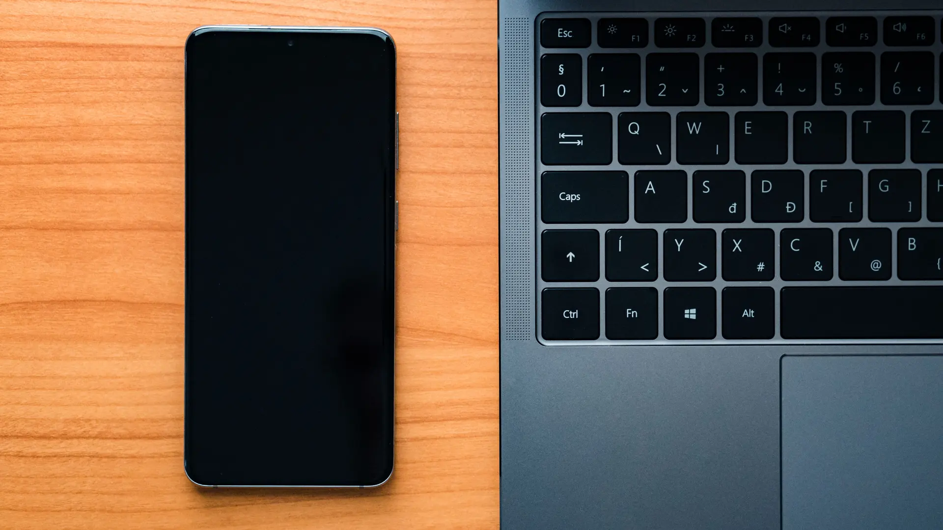 Celular com a tela apagada ao lado do teclado do notebook, numa mesa de madeira minimalista