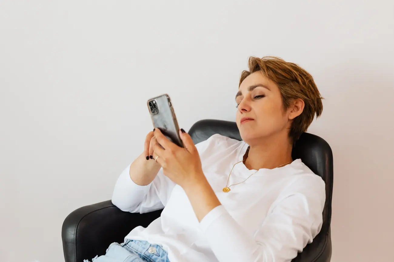 Mulher sentada olhando para a tela do celular, fundo claro e neutro