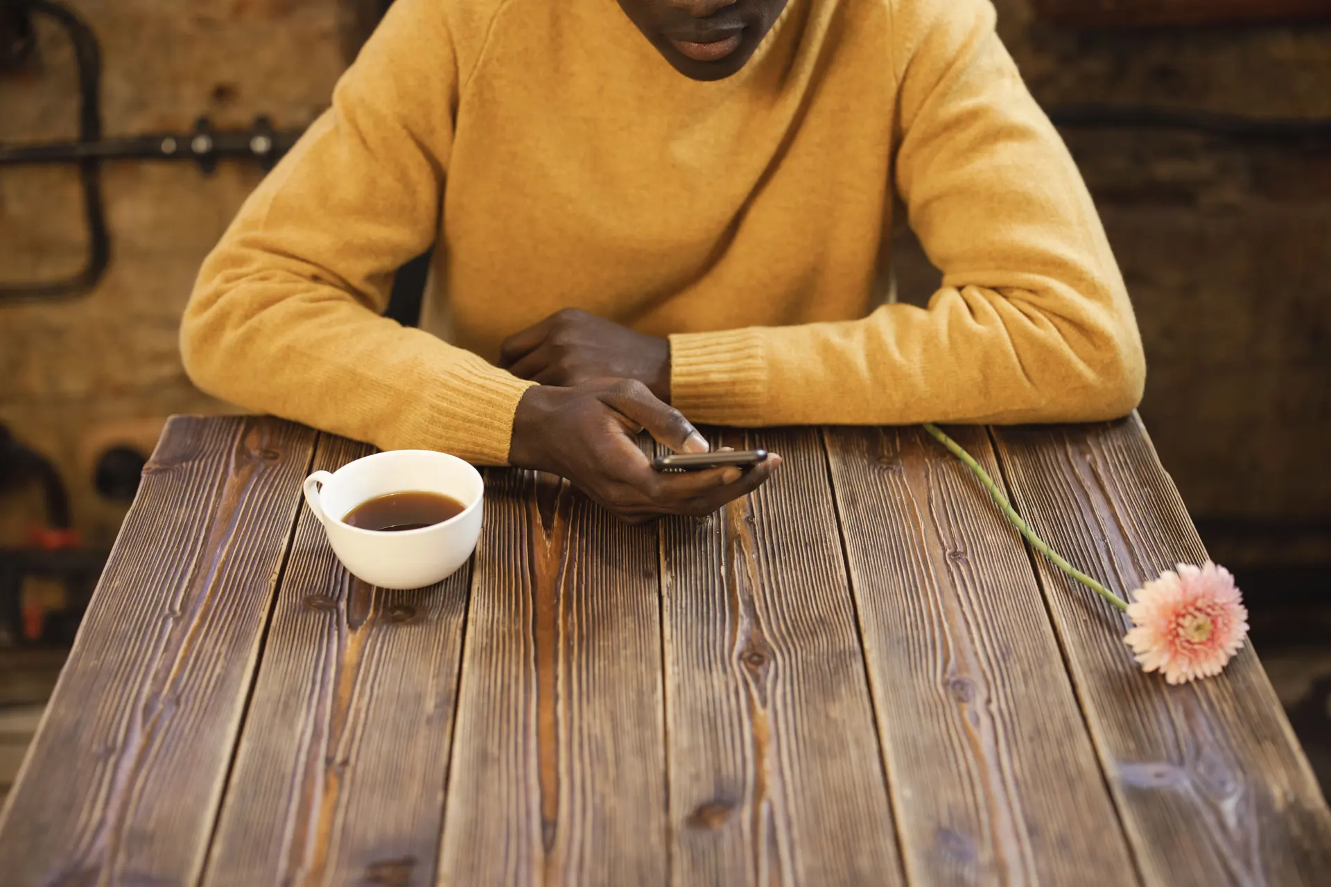 Pessoa consultando o celular em uma mesa de madeira, com café e flor ao lado