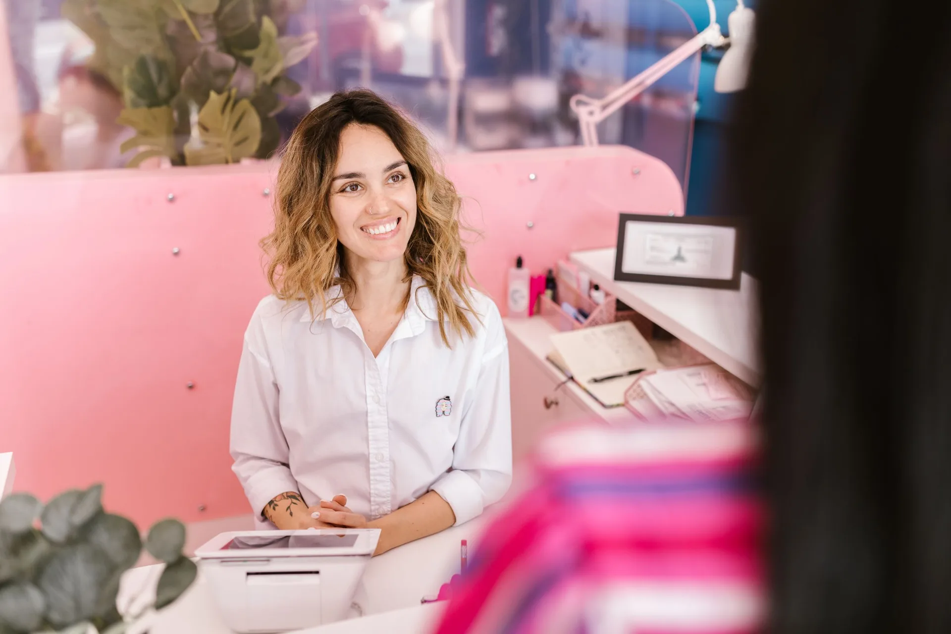 Mulher sorrindo na recepção de um salão de beleza, com um tablet e o caderno de agenda sobre o balcão