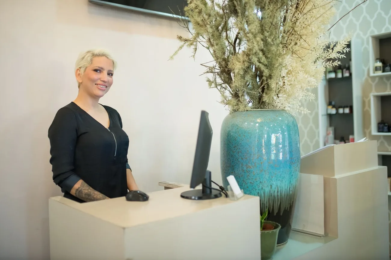 Mulher sorrindo atrás do balcão da recepção de um salão de beleza, ao lado do monitor do computador