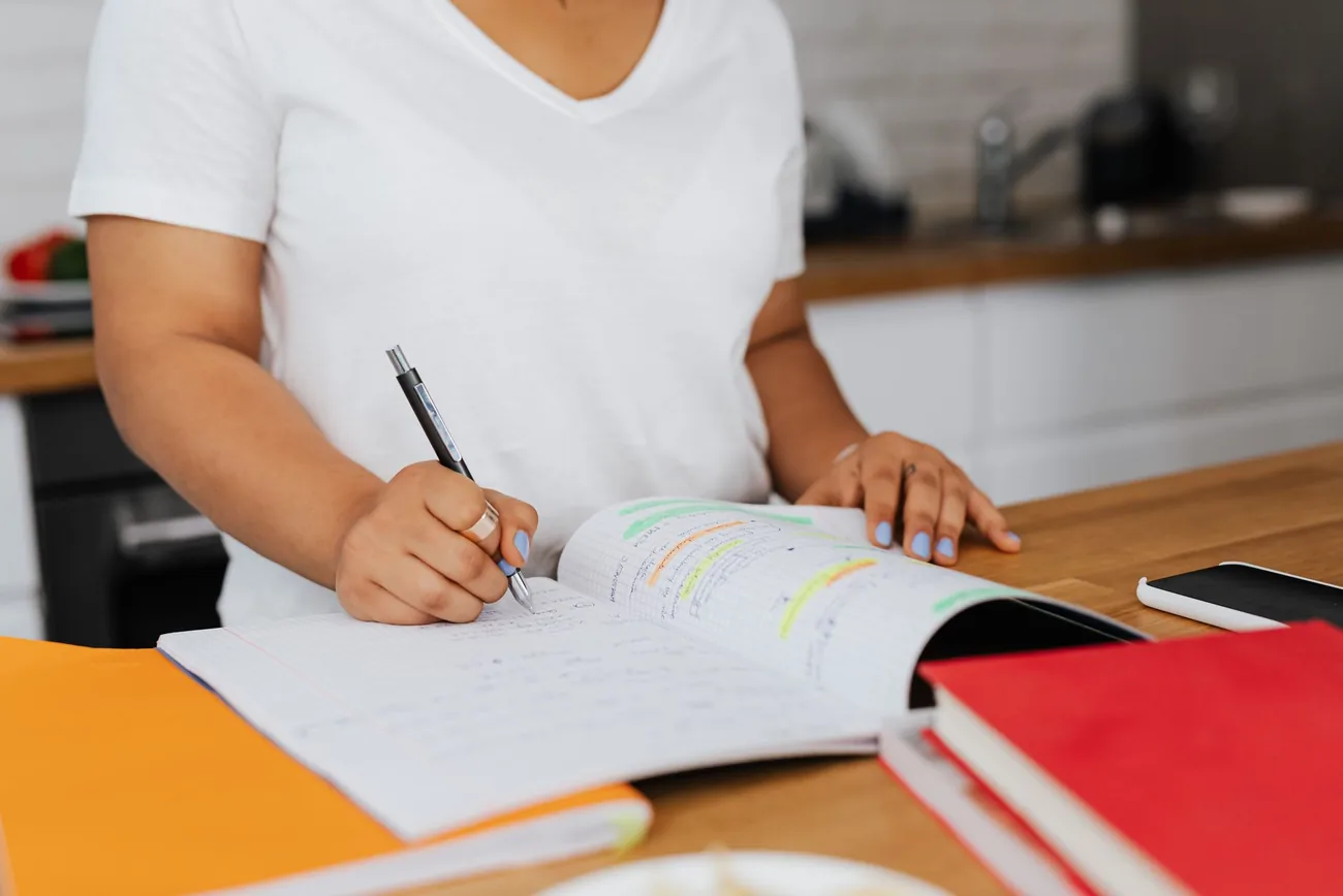 Pessoa escrevendo num caderno aberto sobre a mesa, com o celular ao lado