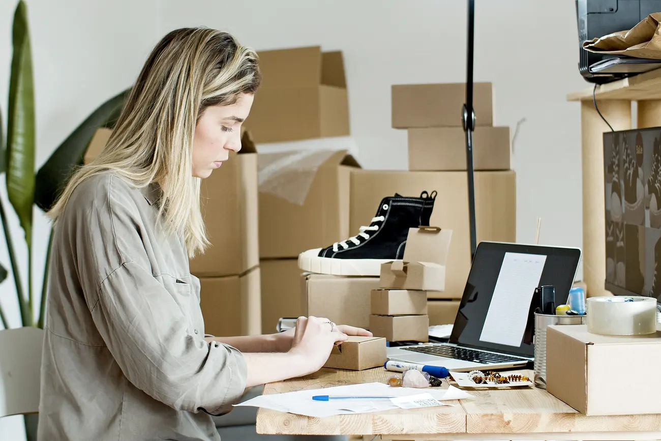Dona de loja embalando um pedido com cuidado ao lado do notebook